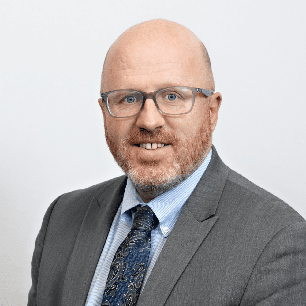 Headshot of Robbie Thompson from the shoulders up, smiling with glasses, in a gray with no hair